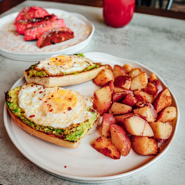 Avocado toast with fried eggs and roasted potatoes, with pink French toast in the background.