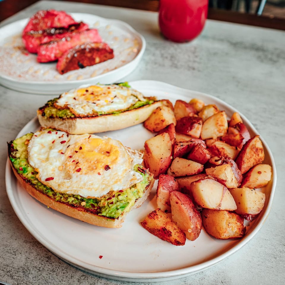 Avocado toast with fried eggs and roasted potatoes, with pink French toast in the background.