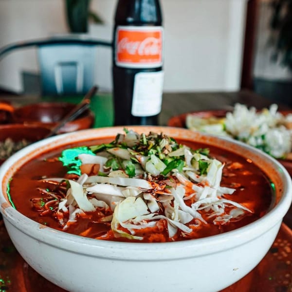 A bowl of red pozole with shredded cabbage and cilantro, with a Coca-Cola bottle in the background.