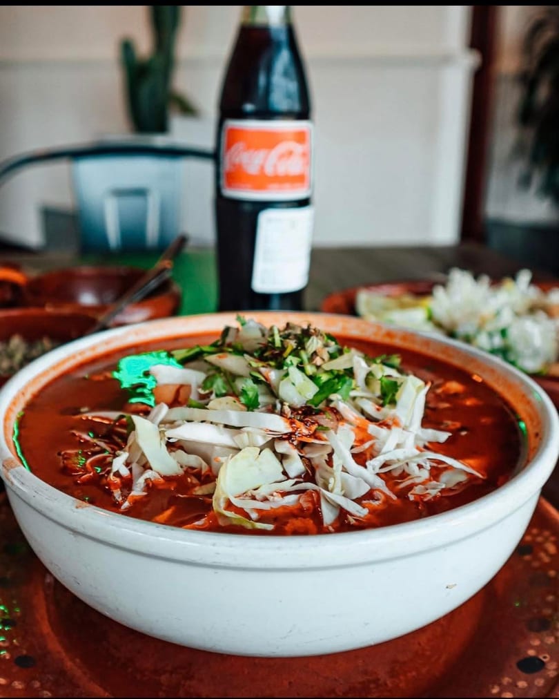 A bowl of red pozole with shredded cabbage and cilantro, with a Coca-Cola bottle in the background.