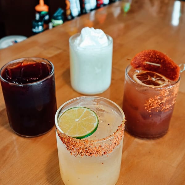 Four colorful cocktails, including a margarita with a lime, on a wooden bar.