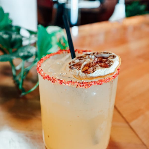 A light-colored cocktail with a red-rimmed glass, straw, and dried citrus slice on a wooden table.