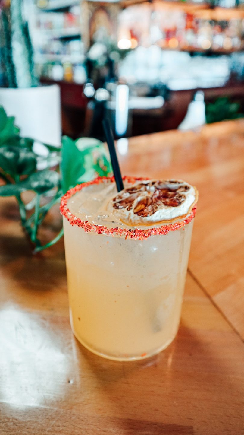 A light-colored cocktail with a red-rimmed glass, straw, and dried citrus slice on a wooden table.